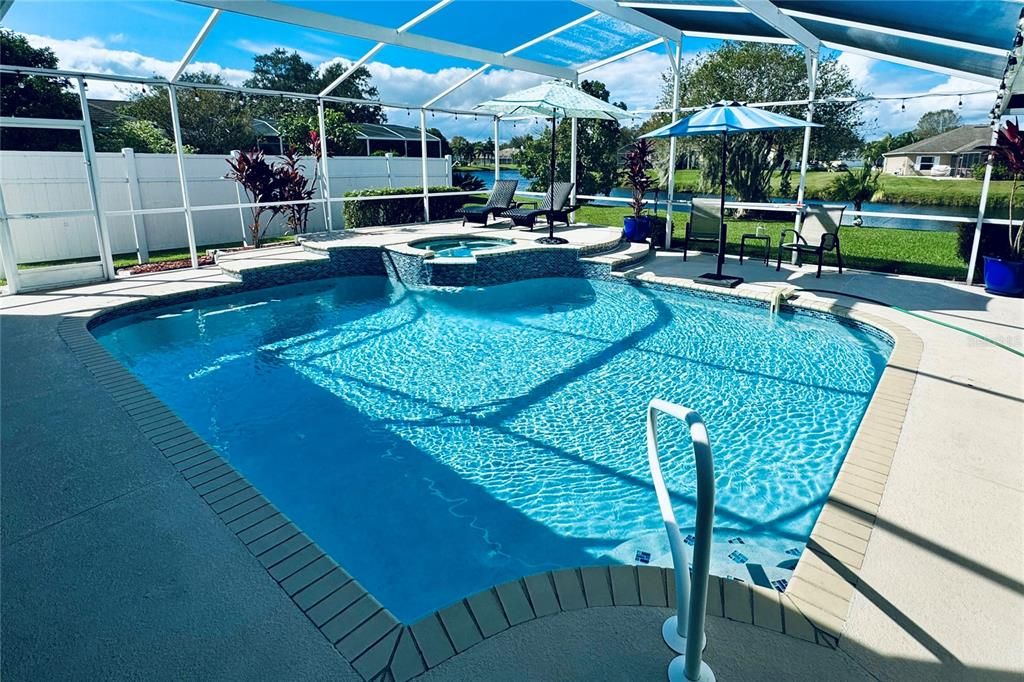 This beautiful screened-in pool area offers a resort-like setting with a sparkling pool and an elevated spillover spa, perfect for relaxation and entertainment. The pool’s design features a spacious deck with ample room for lounging and dining, while the raised spa cascades into the pool, creating a soothing waterfall effect. Beyond the pool, enjoy a tranquil view of the nearby pond, providing a peaceful backdrop to this outdoor oasis. The surrounding landscaping and privacy screens add an extra layer of seclusion. In the evenings, string lights around the enclosure create a warm and inviting ambiance, transforming the space into a serene retreat under the stars. This outdoor area is ideal for both daytime enjoyment and evening relaxation, combining elegance with a touch of nature.