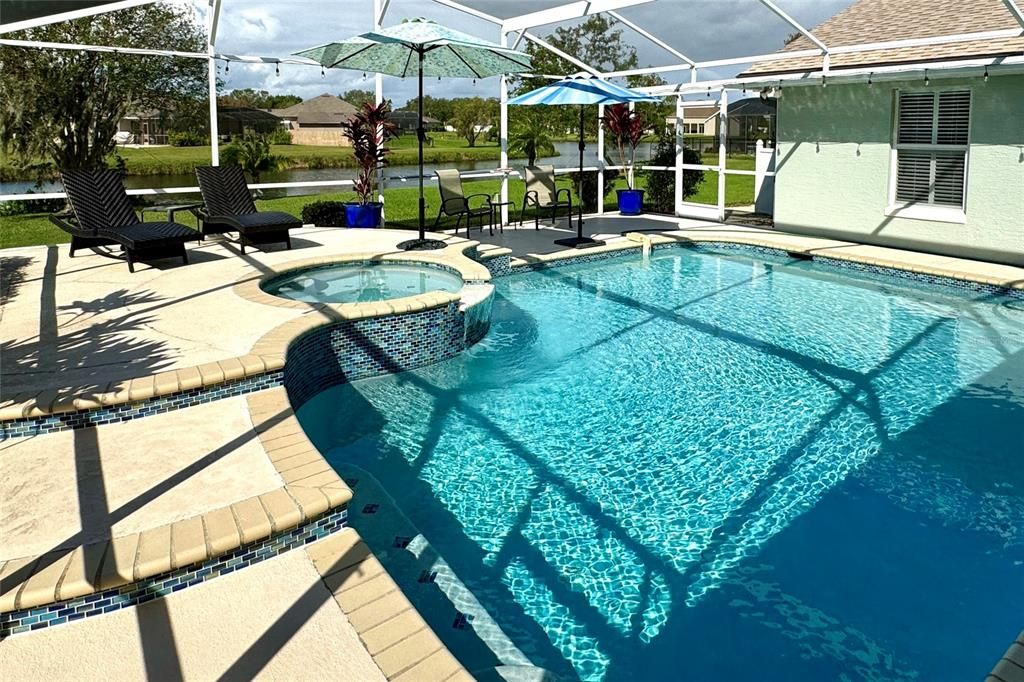 This beautiful screened-in pool area offers a resort-like setting with a sparkling pool and an elevated spillover spa, perfect for relaxation and entertainment. The pool’s design features a spacious deck with ample room for lounging and dining, while the raised spa cascades into the pool, creating a soothing waterfall effect. Beyond the pool, enjoy a tranquil view of the nearby pond, providing a peaceful backdrop to this outdoor oasis. The surrounding landscaping and privacy screens add an extra layer of seclusion. In the evenings, string lights around the enclosure create a warm and inviting ambiance, transforming the space into a serene retreat under the stars. This outdoor area is ideal for both daytime enjoyment and evening relaxation, combining elegance with a touch of nature.