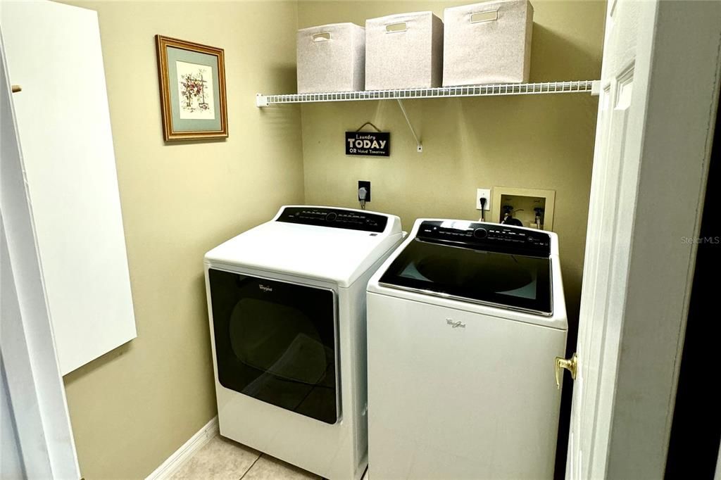 This well-appointed laundry room offers convenience and functionality with an included Whirlpool washer and dryer, easy-maintenance tile flooring, a storage shelf above the appliances, and a built-in fold-down ironing board wired for an iron. Designed to streamline daily routines, this utility space is both stylish and efficient.