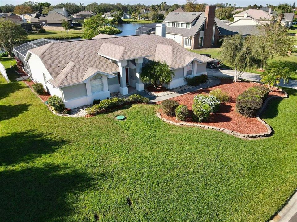 This aerial view showcases a beautifully landscaped single-story home with a fresh roof (replaced in 2022), surrounded by manicured greenery and mulch beds accented with stone edging. The property backs onto a peaceful pond, creating a scenic and serene setting in a spacious neighborhood.