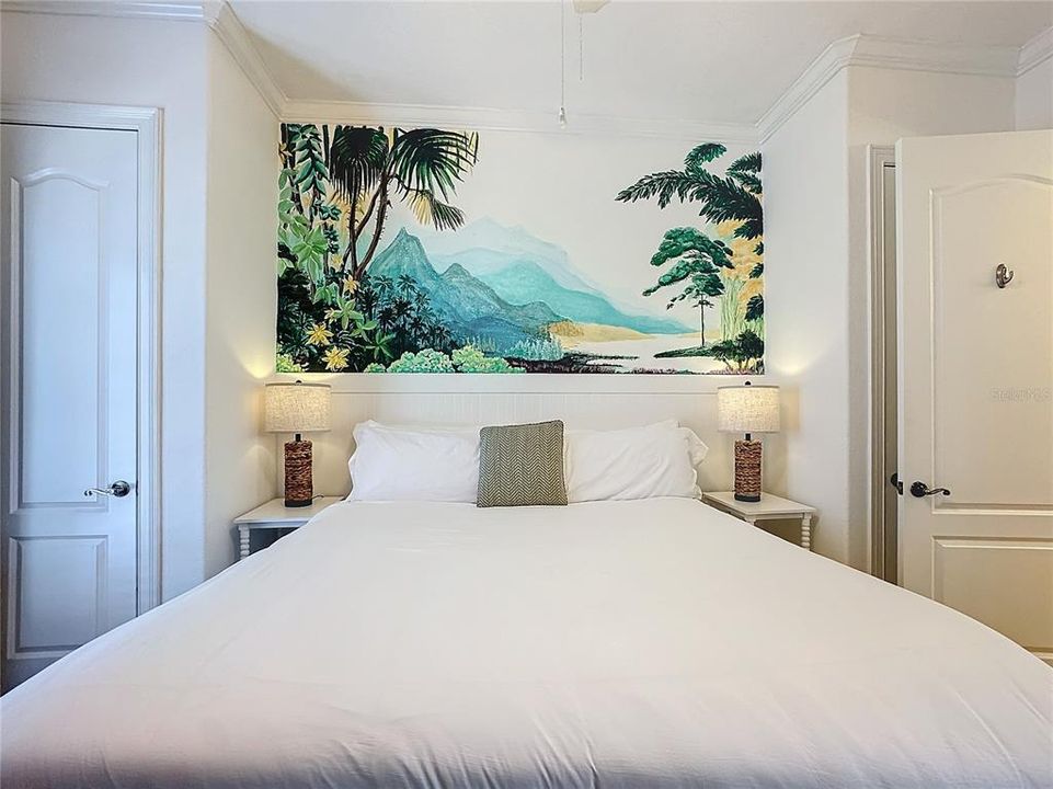 The bedroom is a tranquil escape, where a stunning hand-painted mural serves as the room's centerpiece, stretching gracefully above the bed. The mural depicts a vibrant tropical scene, with lush greenery, swaying palm trees, and distant, misty mountains rising in the background.