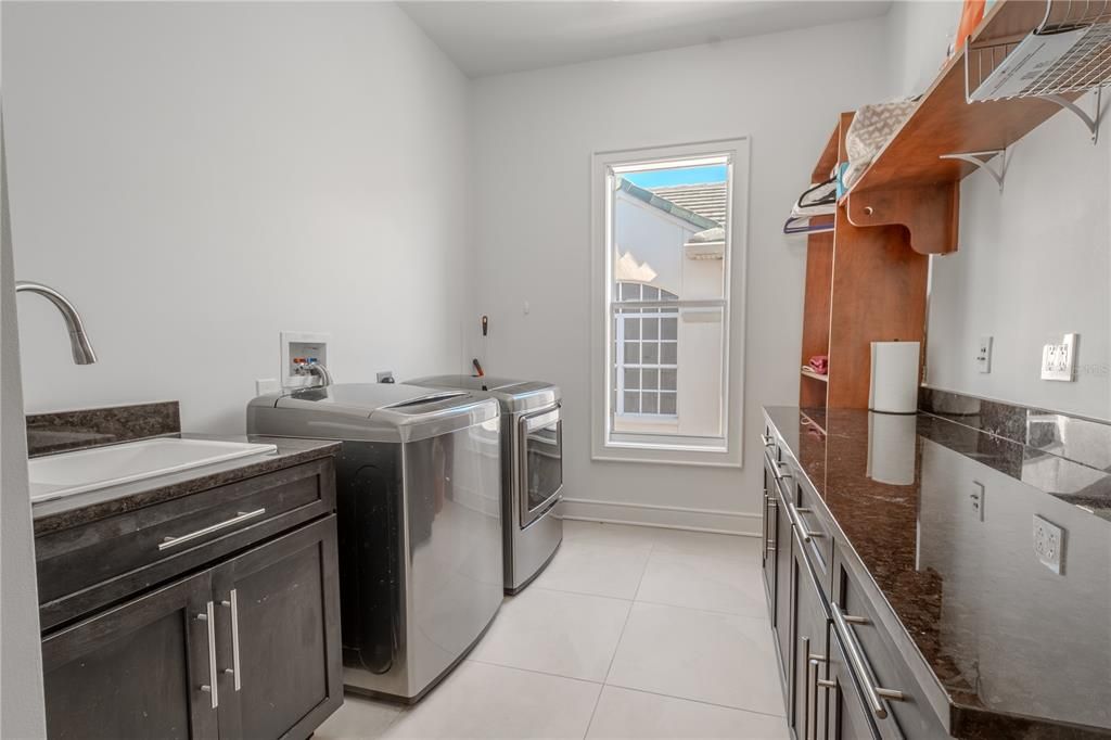 The laundry room features a utility sink with storage, a washer and dryer, shelving, and built-in cabinetry and draws with granite counter top.