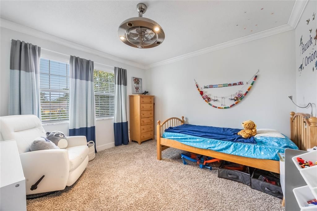 View onto bedroom3, with dual windows on the left and a twin bed located in the middle of the image. There is a stainless steel ceiling fan mounted to the ceiling.