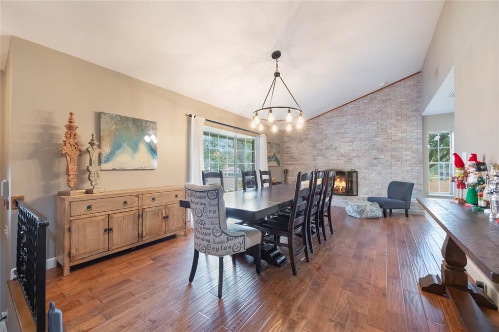 This photo depicts a dining area with a warm and inviting ambiance. The room features a vaulted ceiling with a light beige tone and a rustic chandelier with exposed bulbs hanging above a long, dark wooden dining table surrounded by black chairs. A single chair with a patterned fabric design stands out among the others. To the left of the table, there is a wooden sideboard with decorative carvings and a modern abstract painting mounted above it.The flooring is rich hardwood, adding warmth to the space. In the background, a floor-to-ceiling whitewashed brick wall features a fireplace with a cozy fire burning inside, flanked by large windows with white curtains that let in natural light. On the right, a small sitting area includes a gray upholstered chair and a decorative pillow. A wooden console table is placed against the far-right wall