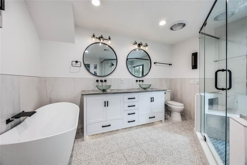 This photo looks into the en-suite primary bathroom. On the left is a standalone tub, in the middle of the image is a large dual sink vanity with bowl sinks sitting on top of the counter and two round mirrors handing above. To the fight is the stall shower with glass enclosure.