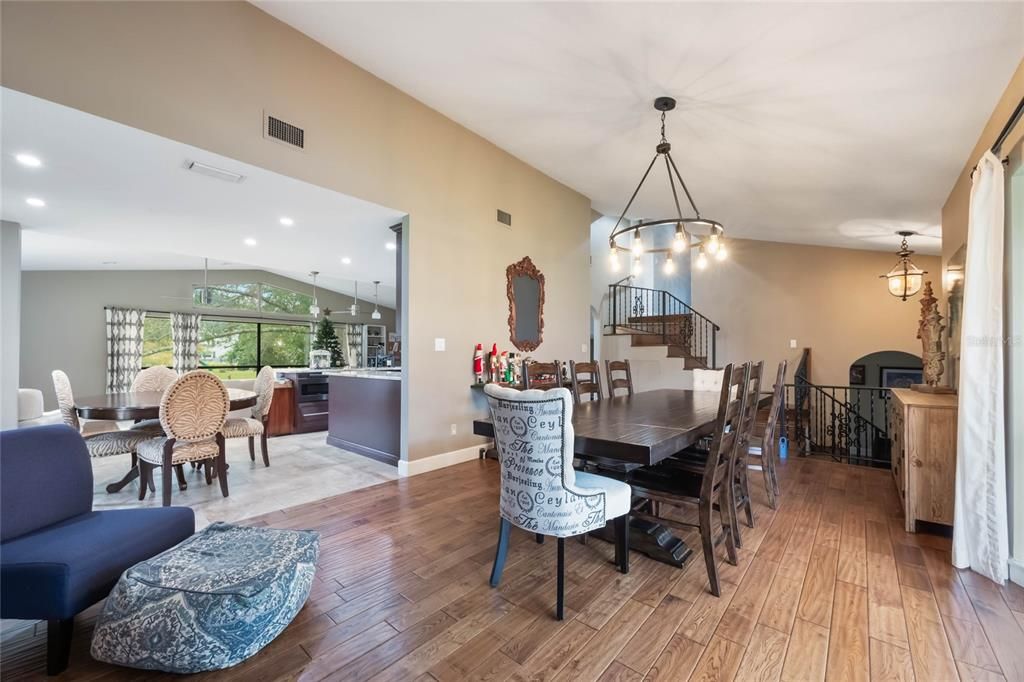 This image  showcases a dining area that opens into a living space and kitchen. The dining area features a long, dark wooden table surrounded by matching chairs, with a single accent chair upholstered in patterned fabric at one end. Above the table, a rustic chandelier with exposed Edison-style bulbs hangs from the ceiling, casting warm light across the space.In the background, a staircase with black wrought iron railing leads to an upper level, while the walls are painted in neutral tones, adding to the warm and inviting ambiance. On the right, holiday decorations and a mirror adorn a console table. The flooring is rich hardwood, which transitions into lighter tile as the space opens into the kitchen and living area, visible through a large, open archway. The kitchen is equipped with modern appliances and features an island with a granite countertop and barstool seating. The living area beyond includes large windows, allowing ample natural light to flood the space.