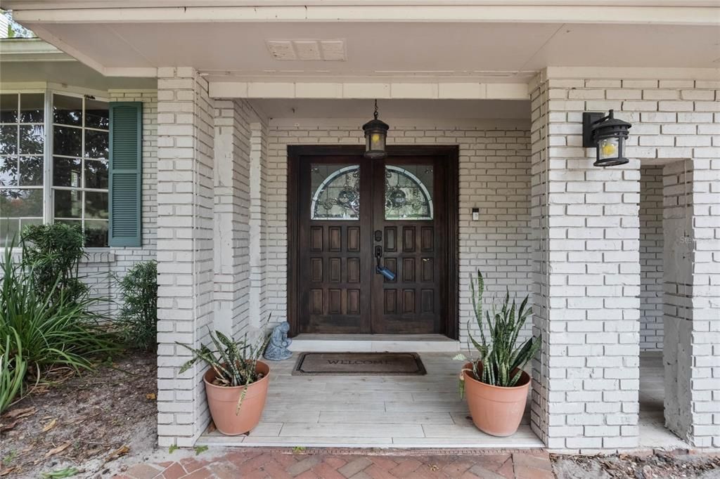 This photo shows the front entrance of a home with a white-painted brick exterior. The entryway features a pair of dark wooden double doors with decorative arched glass inserts. A hanging lantern-style light fixture is centered above the doorway, and matching wall-mounted lights are installed on either side of the entrance. Two terracotta pots with snake plants (Sansevieria) are positioned symmetrically on either side of the door. A beige "Welcome" mat lies on the tiled floor of the small covered porch. To the left, a window with green shutters is partially visible, framed by surrounding greenery. The porch is bordered by a brick-paved walkway.