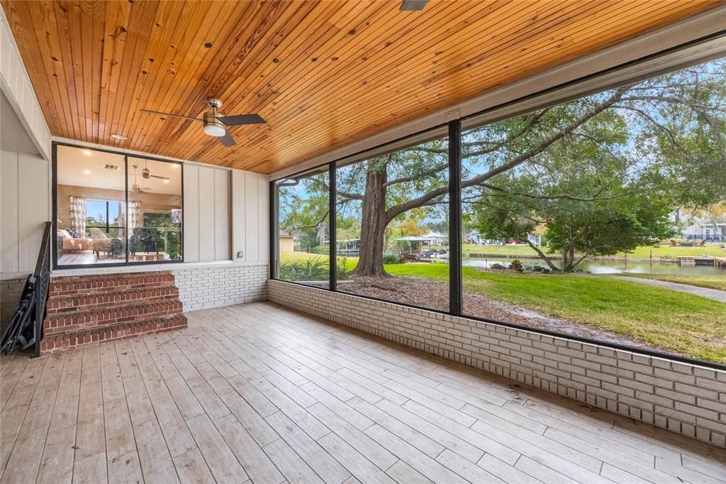 This photo features the back screened patio. In the upper left of the image is a set of stairs that lead into the family room. The ceiling shows off a beautiful wood finish and to the right of the image shows views of the backyard with mature trees and canal that run the length of the property.