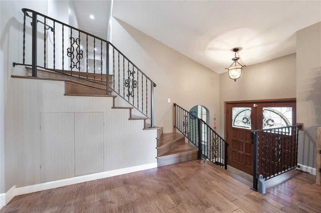 This photo looks from the dinging area toward the beautiful wooden double doors and open stairs leading to the 2nd level of the home. The stairs are lined with wrot iron railings that lead to a hallway.