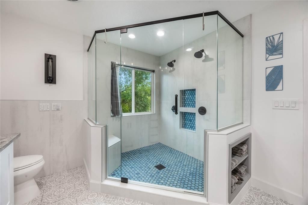 This photo shows the large stall shower in the primary bathroom. The shower has a light grey tile on the walls with a blue mosiac tile floor. There are also two shower heads with two separate controls.