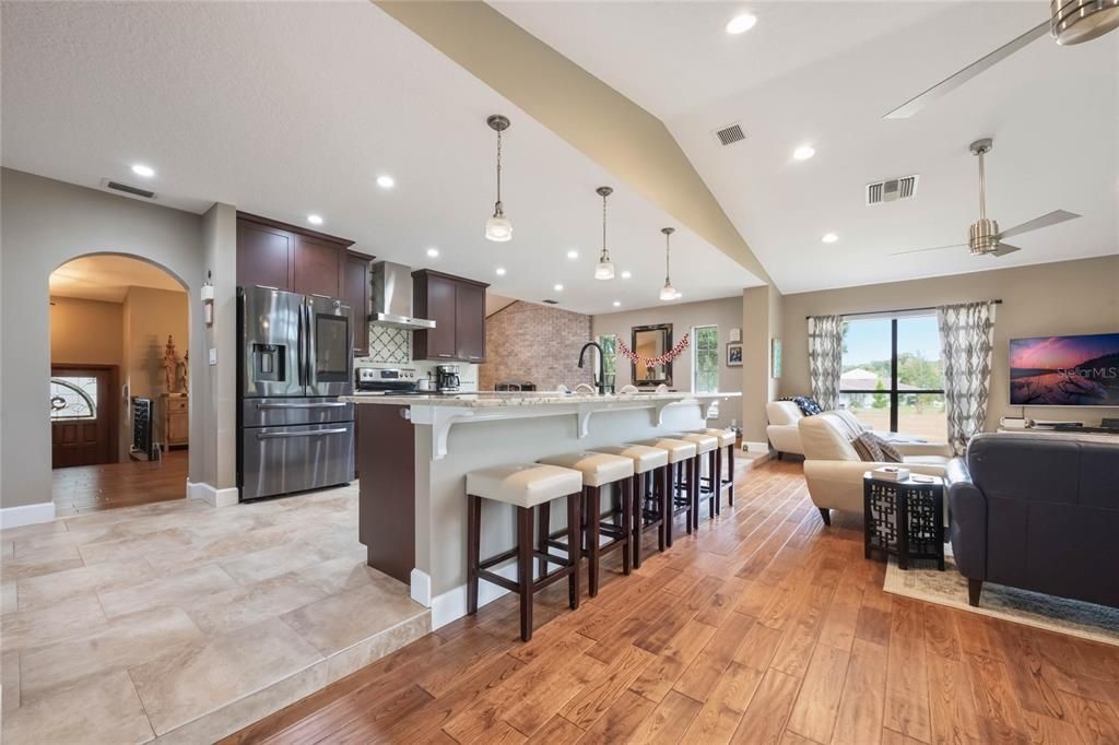 View into the kitchen area from the family room showing the breakfast bar in the middle and dark wood cabinets in tbe back ground with a stainless refrigerator.