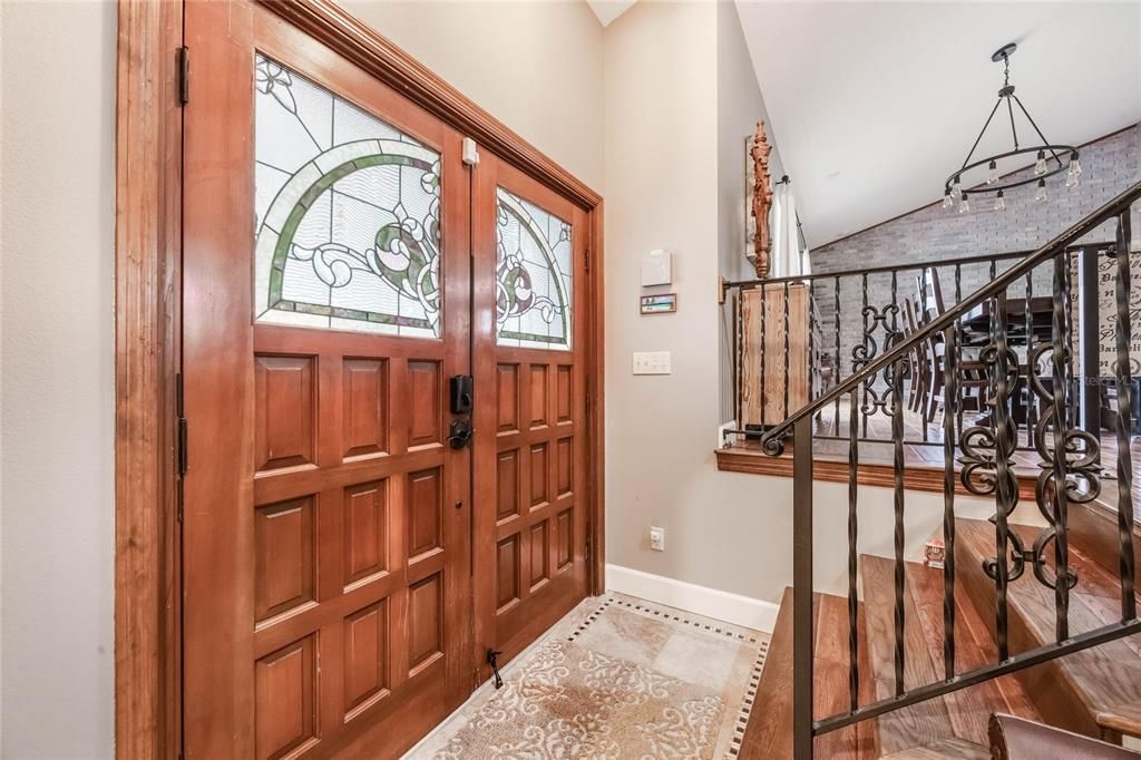 This photo shows the interior entryway of a home. The focal point is a set of wooden double doors with intricate carved panels and decorative stained-glass inserts, featuring floral and geometric patterns. A tiled entryway with a patterned area rug adds warmth to the space. To the right, a staircase with dark wood steps and an ornate wrought-iron railing leads up to an elevated area. In the background, a dining space is partially visible, with a brick accent wall and a chandelier featuring exposed bulb fixtures. The neutral walls and natural wood tones create a welcoming and classic aesthetic.