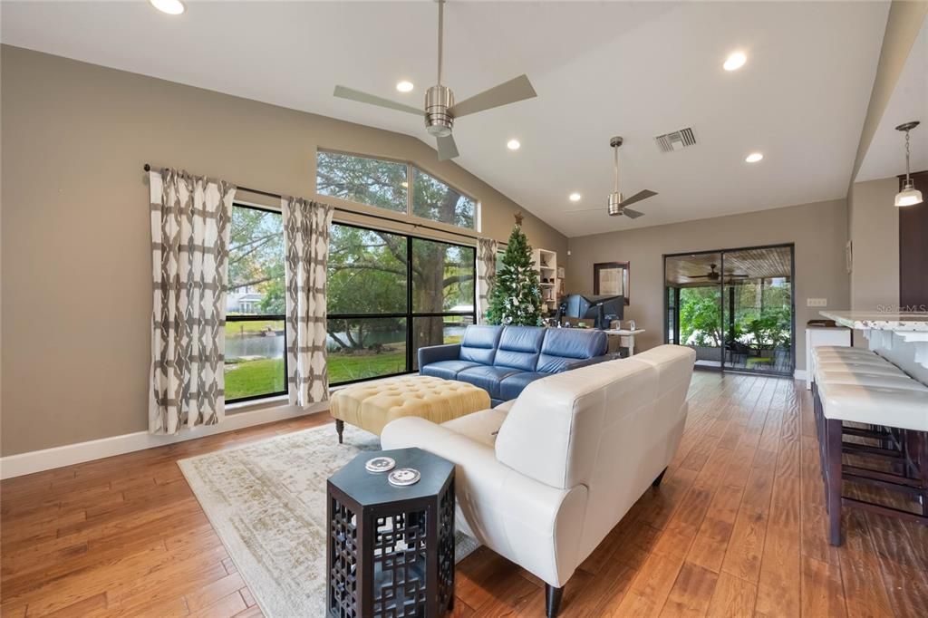 View of the family room from the breakfast nook with a light sofa on the right and brown leather sofa toward the end of the room. A very large set of windows on the left of the picture showing a mature tree in the yard.