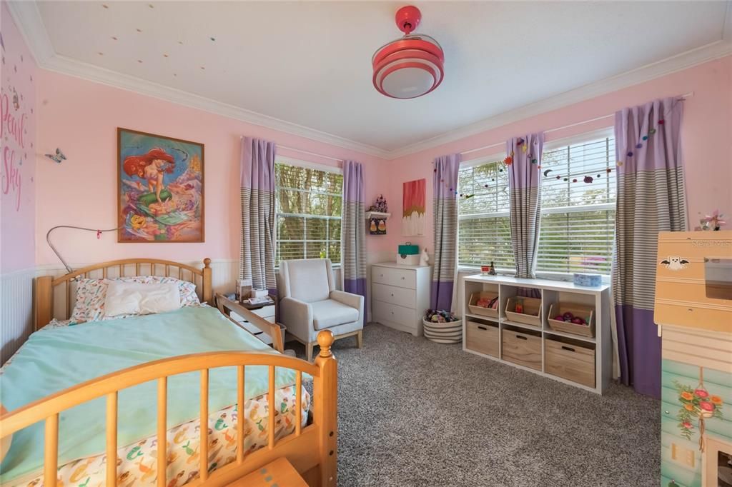 View into the second bedroom that features light pink walls, and a twin bed to the left. in the center of the photo is a window on the back wall of the room and to the right are double windows. There is a dark pink ceiling fan hanging from the ceiling with invisiable fan blades.