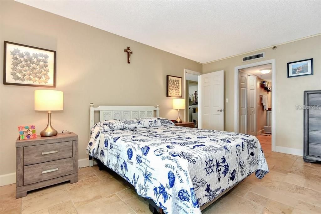 Spacious primary bedroom with travertine floors, coastal decor, and a stunning ocean view from the adjacent living room.