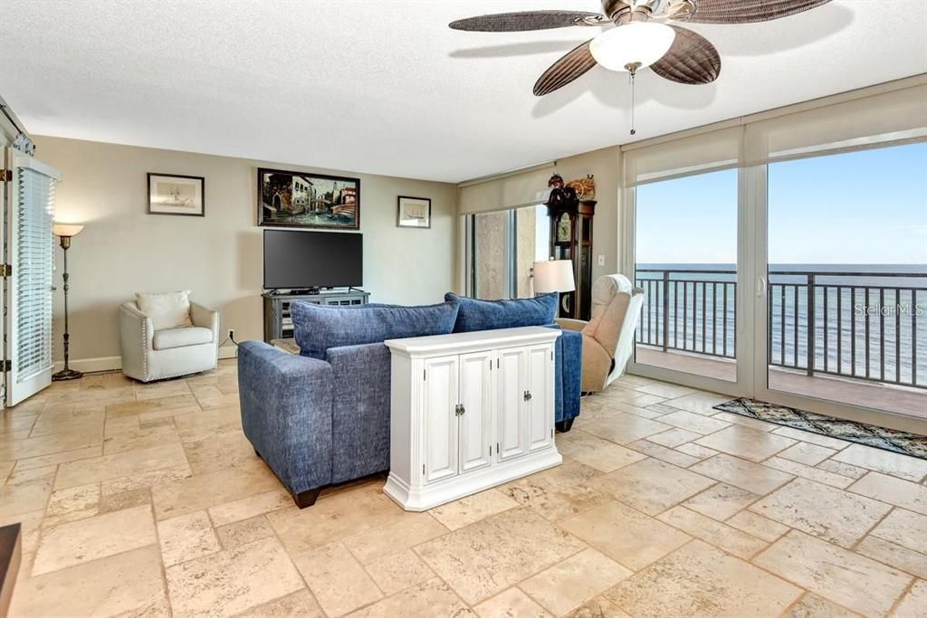 Spacious living room with travertine flooring, a cozy atmosphere, and direct access to the primary suite for seamless functionality.