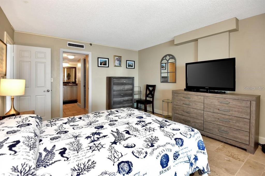 Spacious primary bedroom with travertine floors, coastal decor, and a stunning ocean view from the adjacent living room.