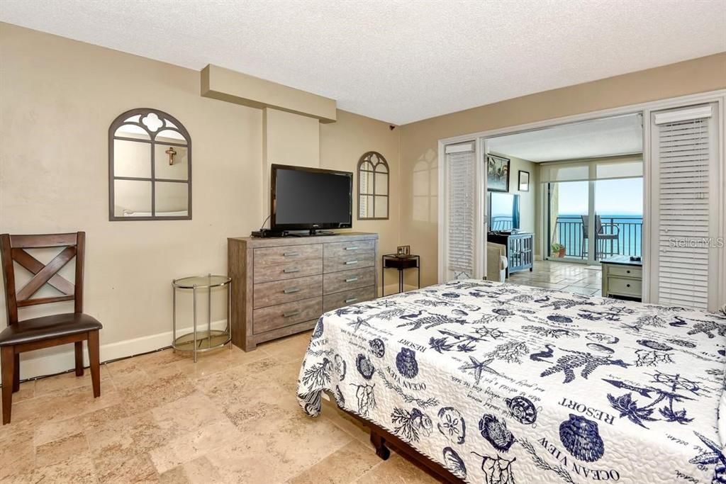 Spacious primary bedroom with travertine floors, coastal decor, and a stunning ocean view from the adjacent living room.