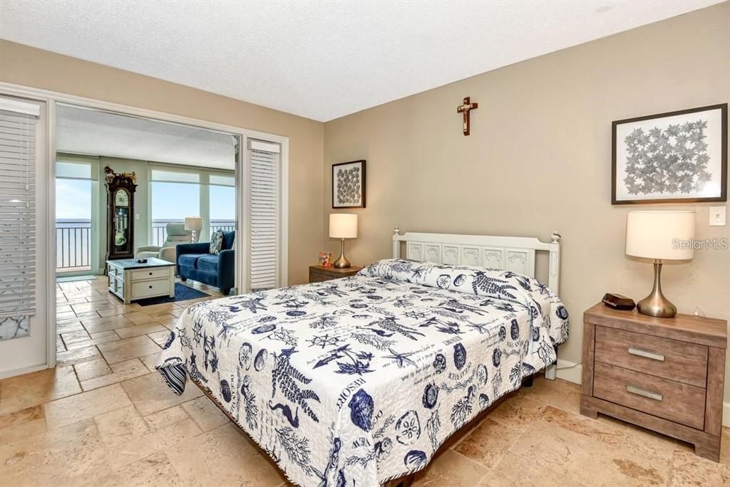 Spacious primary bedroom with travertine floors, coastal decor, and a stunning ocean view from the adjacent living room.