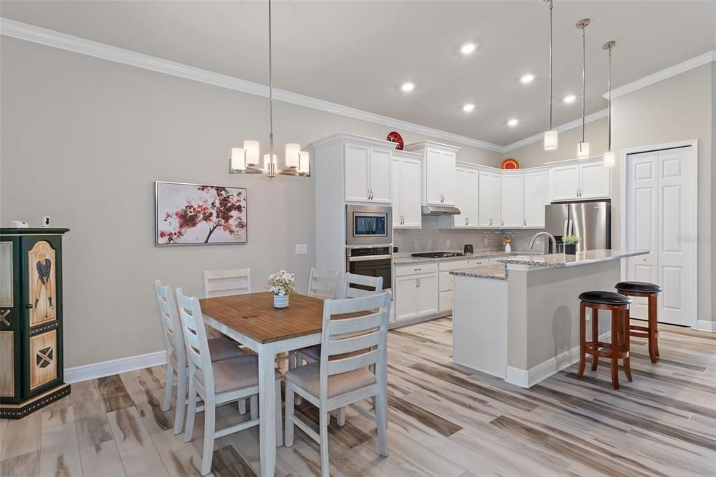 View from between the great room and dining area, looking across into kitchen. Additional features include crown molding, staggered upper cabinets with crown topper, pendant lights, stainless appliances. The pantry is accessed by the door on the right.