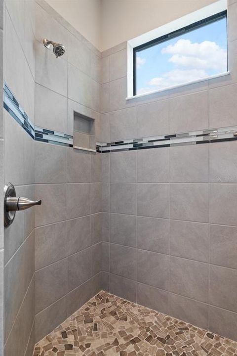 The generous primary bath shower features a shampoo niche, glass tile accent ribbon, high fixed transom window for natural light and privacy, and a smooth pebble floor.