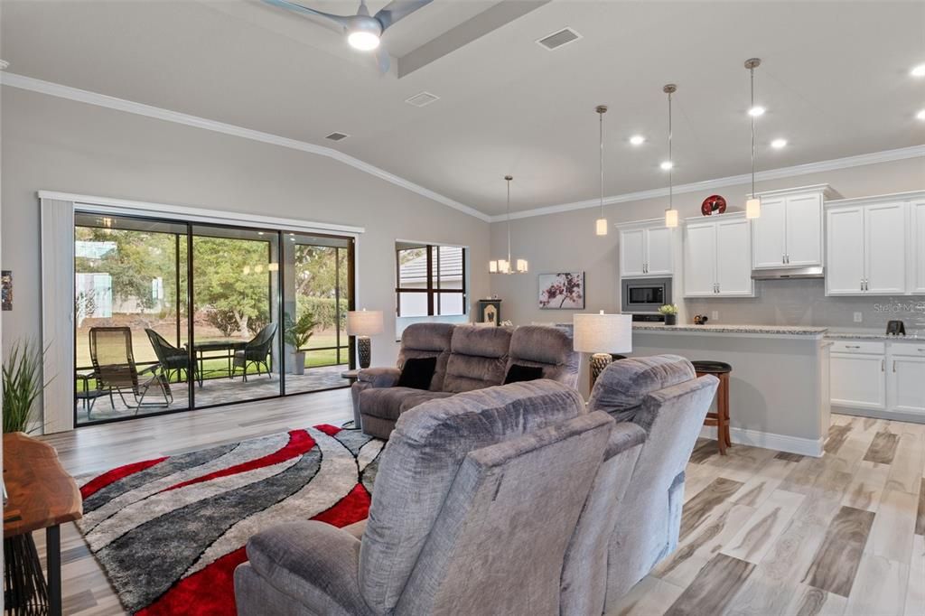 Looking across the great room toward the kitchen and dining nook. Notice additional features such as vertical, pleated accordion blind on sliding door, ceiling fan in great room, pendant lights over breakfast bar, and stylish luxury vinyl plank (LVP) flooring.