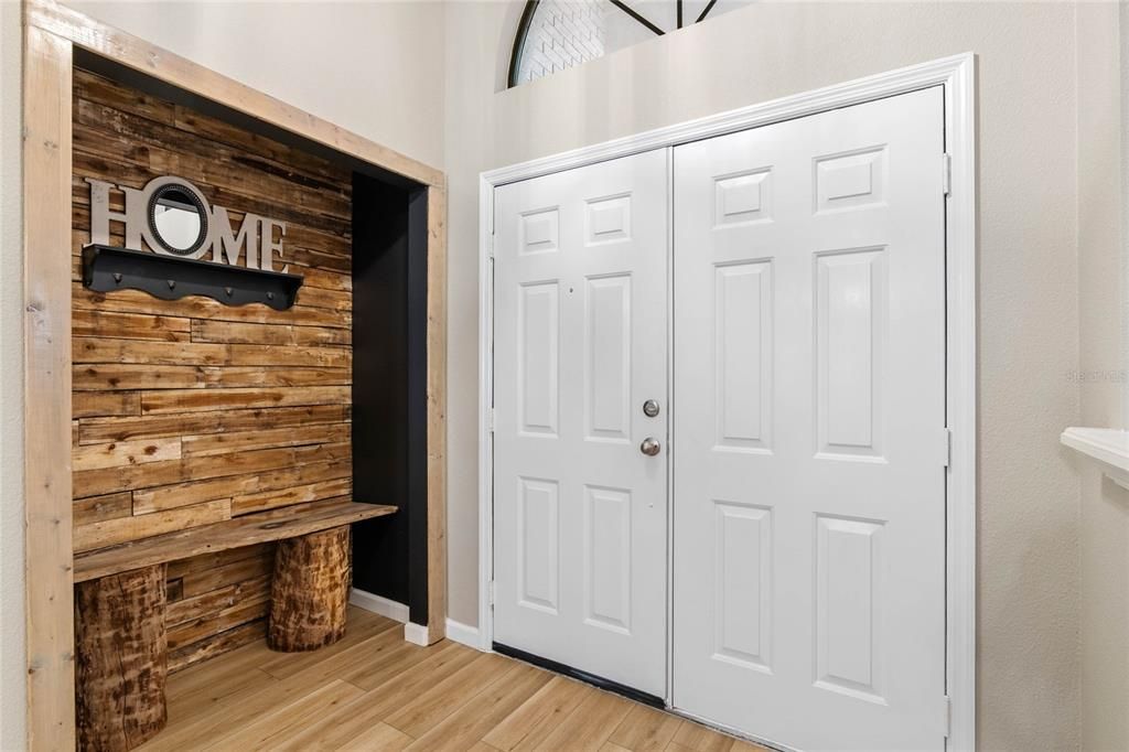 Welcoming entryway featuring double doors with a cozy nook showcasing rustic charm with its wood-paneled accent wall and natural log-style bench. The arched window above the doors allows natural light to flow in, creating an airy and open feel.