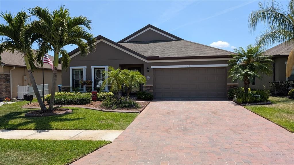 Beautifully landscaped front yard with lush tropical palms, manicured garden beds, and a brick-paver driveway leading to a two-car garage. The home's exterior showcases a warm, neutral color palette with stone accents and inviting curb appeal.